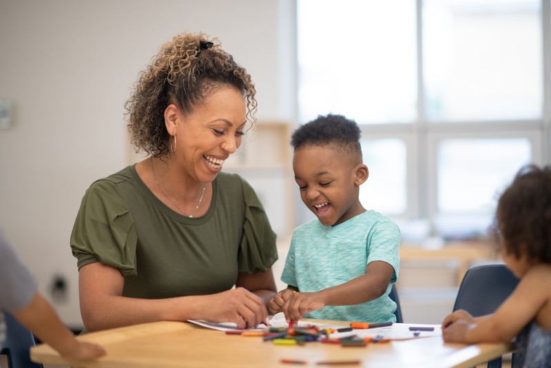 A caregiver shares a joyful coloring moment with a young child at a sunlit table during a naturalistic teaching session.