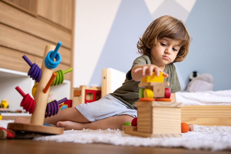 A young child stacks colorful wooden blocks on a cozy bedroom rug during a child-led naturalistic teaching session.