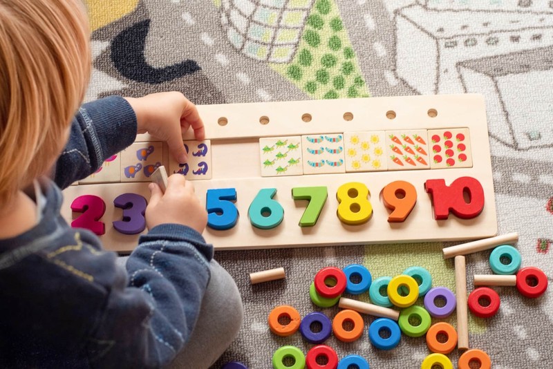 A young child works on a wooden number-matching board with rainbow numerals during a structured ABA teaching task.