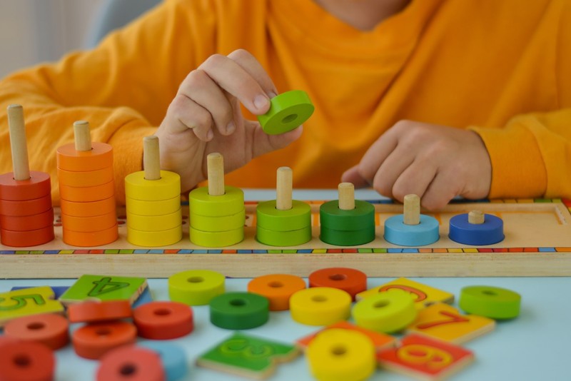 A young child carefully places a green wooden ring onto a stacking peg toy during a structured discrete trial teaching session.