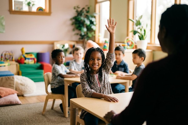 A young African American girl confidently raises her hand during a small-group session at a welcoming ABA therapy center.